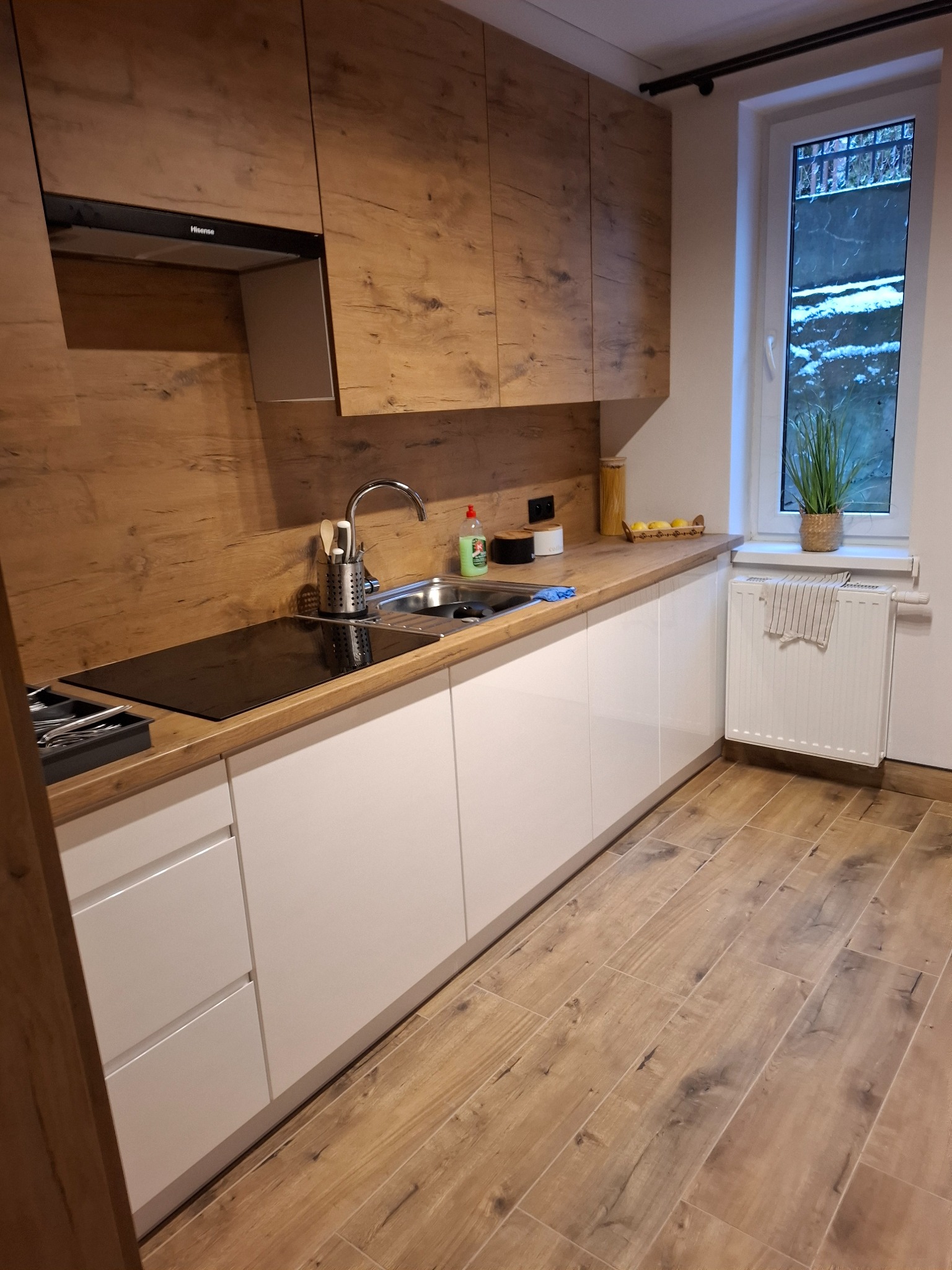 Nowoczesna kuchnia z białymi szafkami dolnymi i górnymi szafkami z drewnopodobnym fornirem, z widocznym okapem Hisense, blatem z płyty drewnopodobnej, płytą indukcyjną i zlewozmywakiem,...