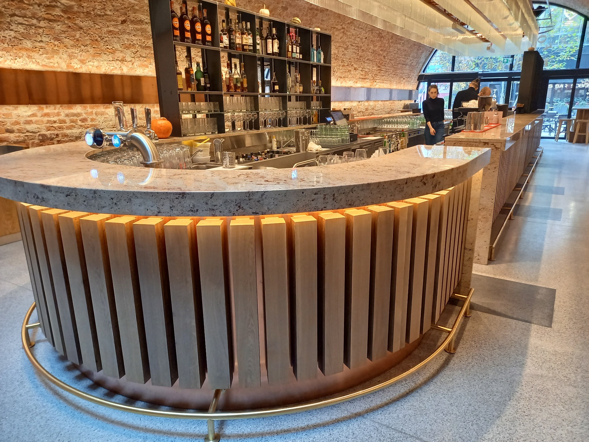 Zaokrąglony bar z blatem z szarego kamienia i drewnianą okładziną z pionowych listew, podświetlonych od dołu, w tle regał z butelkami i szklankami, ceglana ściana i obsługa.