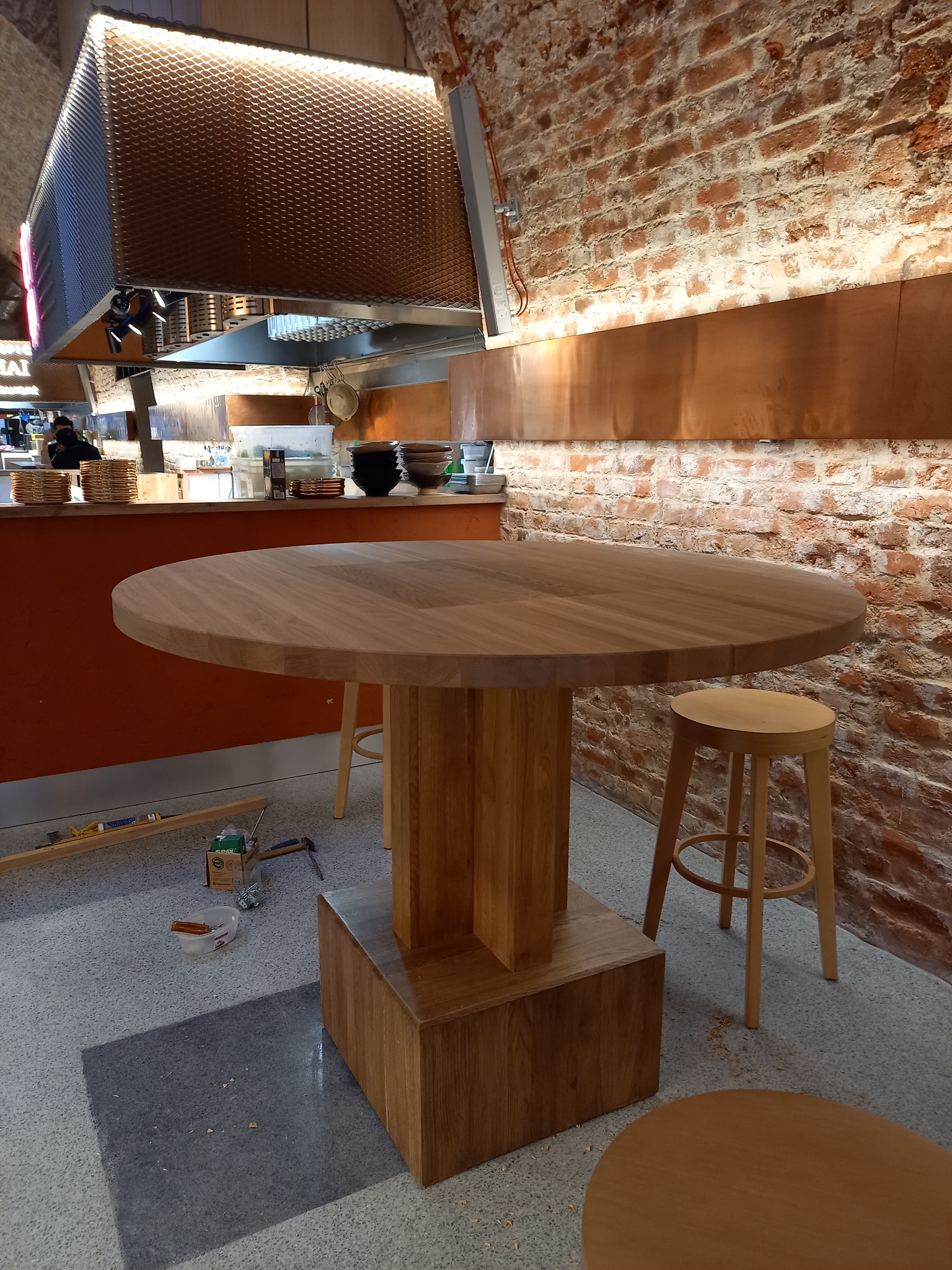Okrągły, surowy blat drewniany na masywnej podstawie w trakcie montażu, widoczne narzędzia i krzesło barowe, tło z cegły.