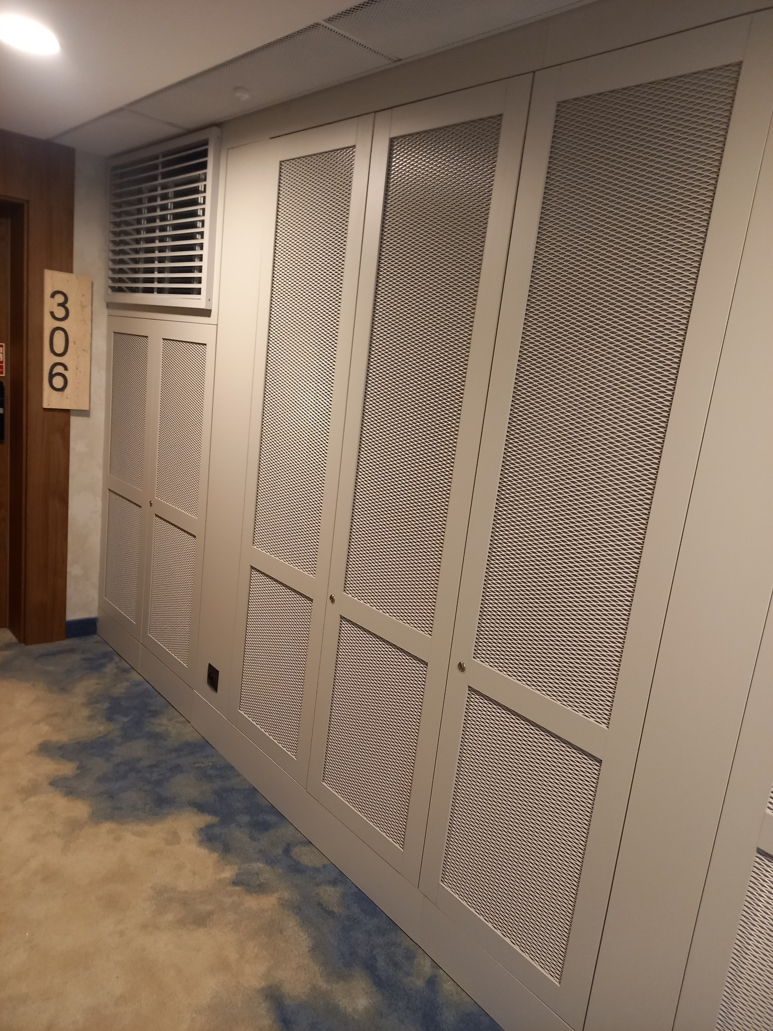 Długi rząd szaf wnękowych z metalową siatką w jasnym korytarzu hotelowym obok numeru pokoju 306.