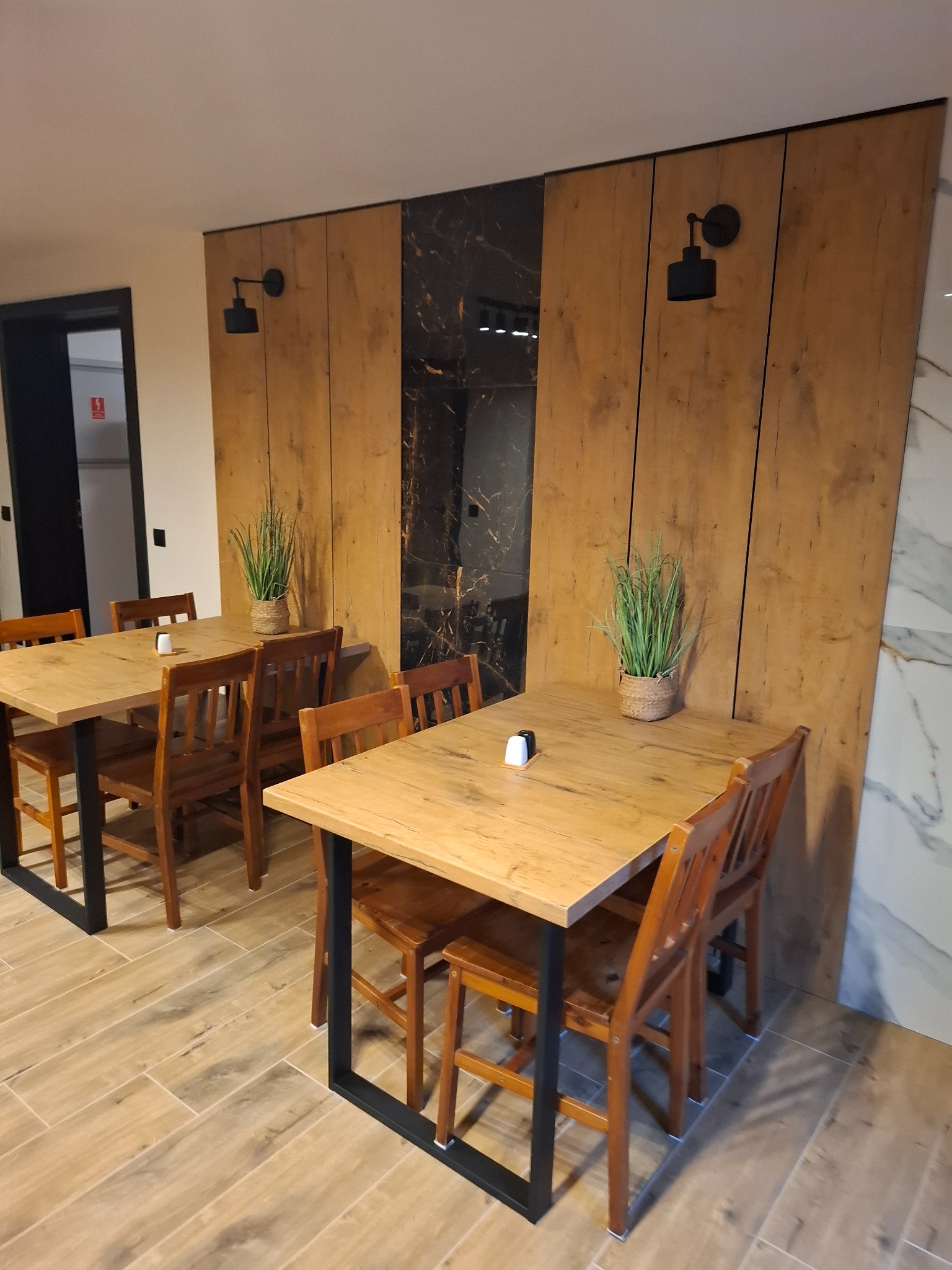 Dwie drewniane stoły z krzesłami na tle ściany z drewnianych paneli, oświetlone czarnymi lampami ściennymi, z roślinami w doniczkach na stołach.