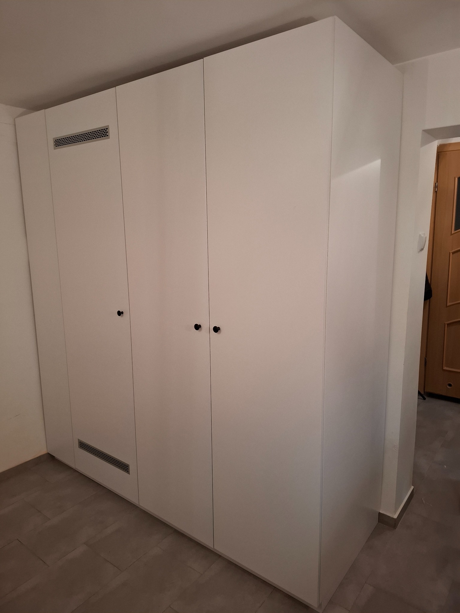 Biała, zabudowana szafa z czterema drzwiami, z czarnymi okrągłymi uchwytami, wbudowana od podłogi do sufitu, z kratkami wentylacyjnymi u góry i dołu, na podłodze z szarych płytek.