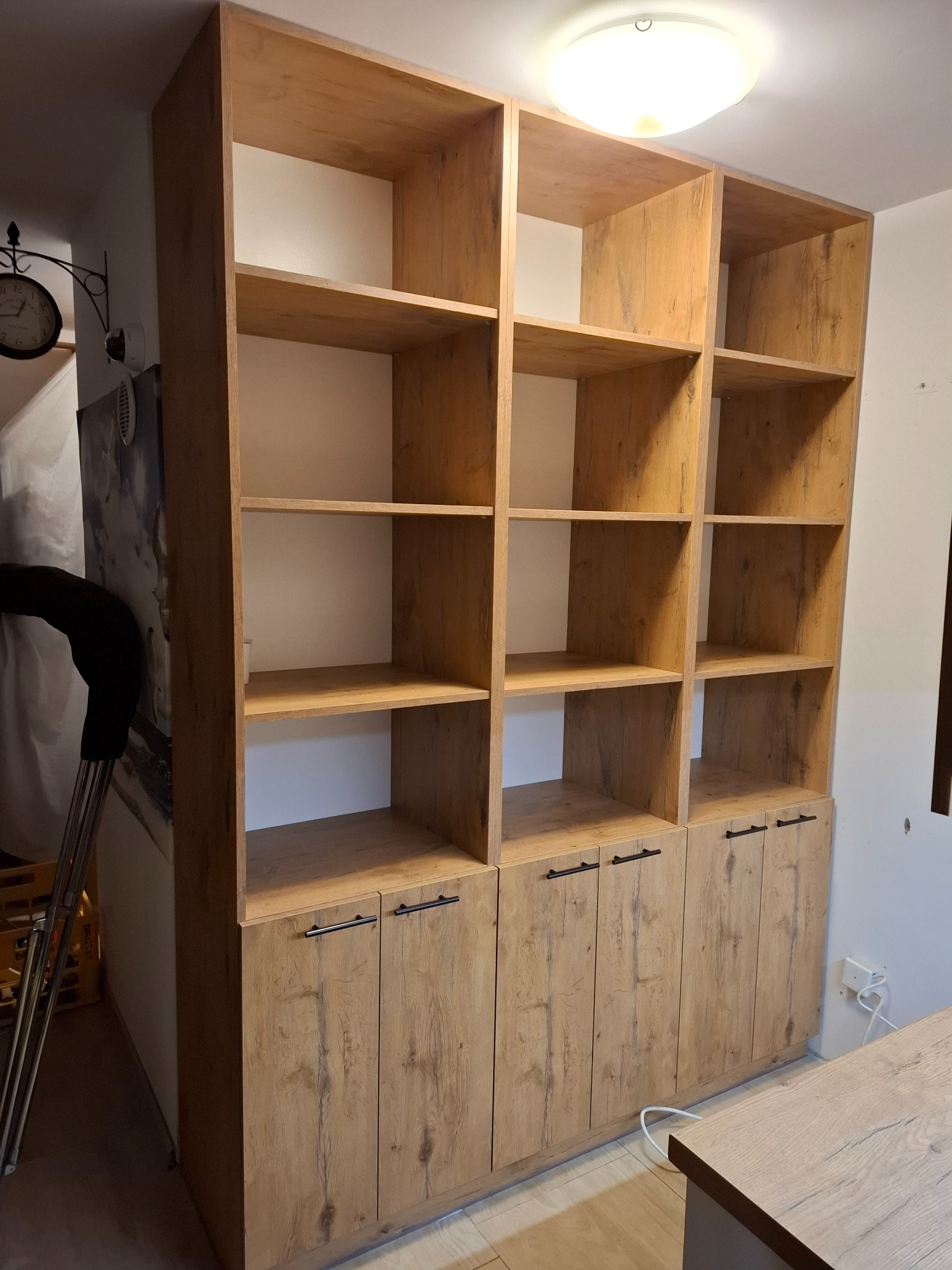 Regał z drewnopodobnej płyty z szafkami w dolnej części, z czarnymi uchwytami, oświetlony lampą sufitową, widoczny fragment ściany z zegarem i obrazem.