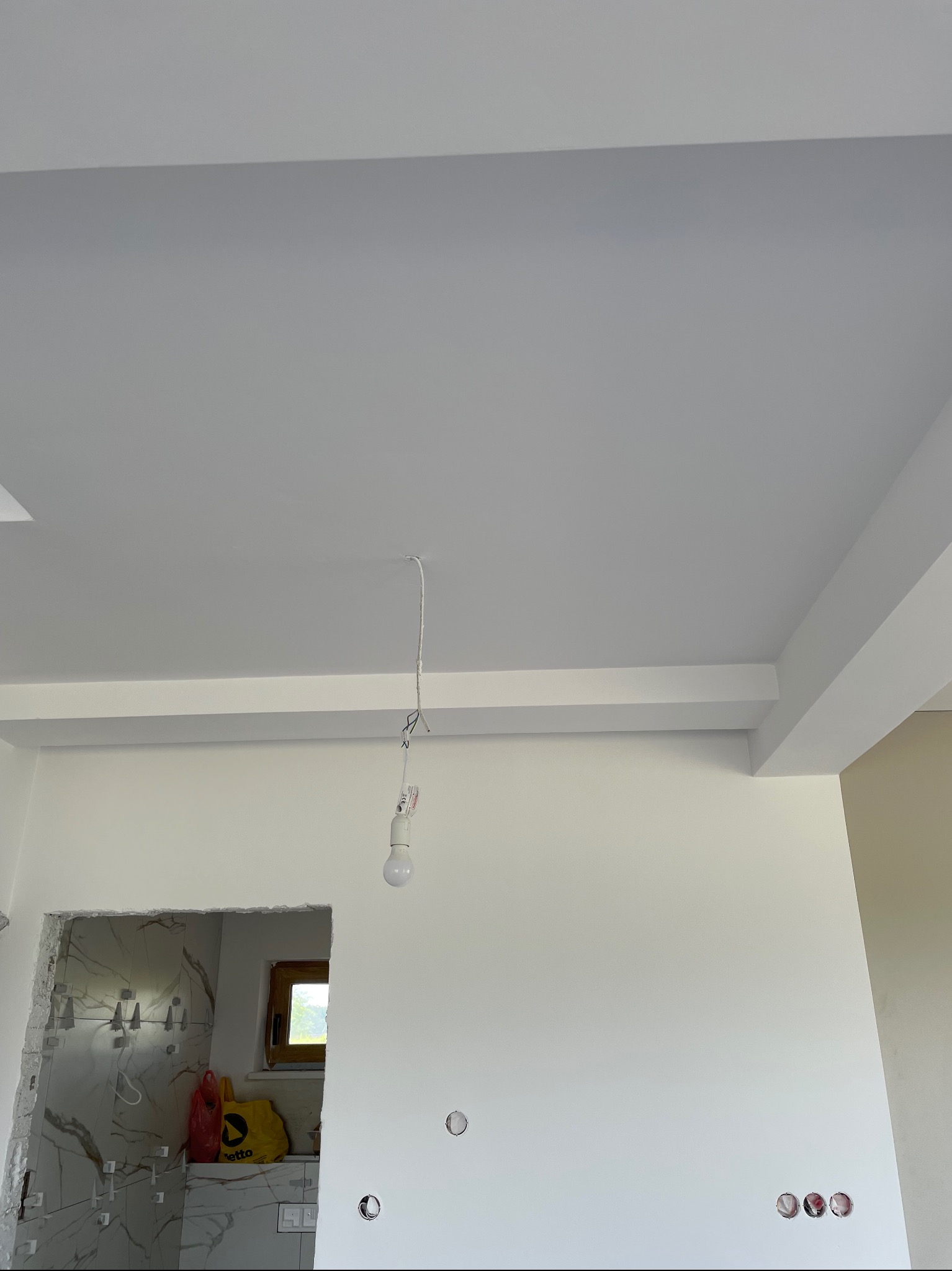 Białe, świeżo otynkowane wnętrze z widocznym sufitem, ścianami i przygotowanym otworem na drzwi do łazienki z marmurowymi płytkami. W tle okno.