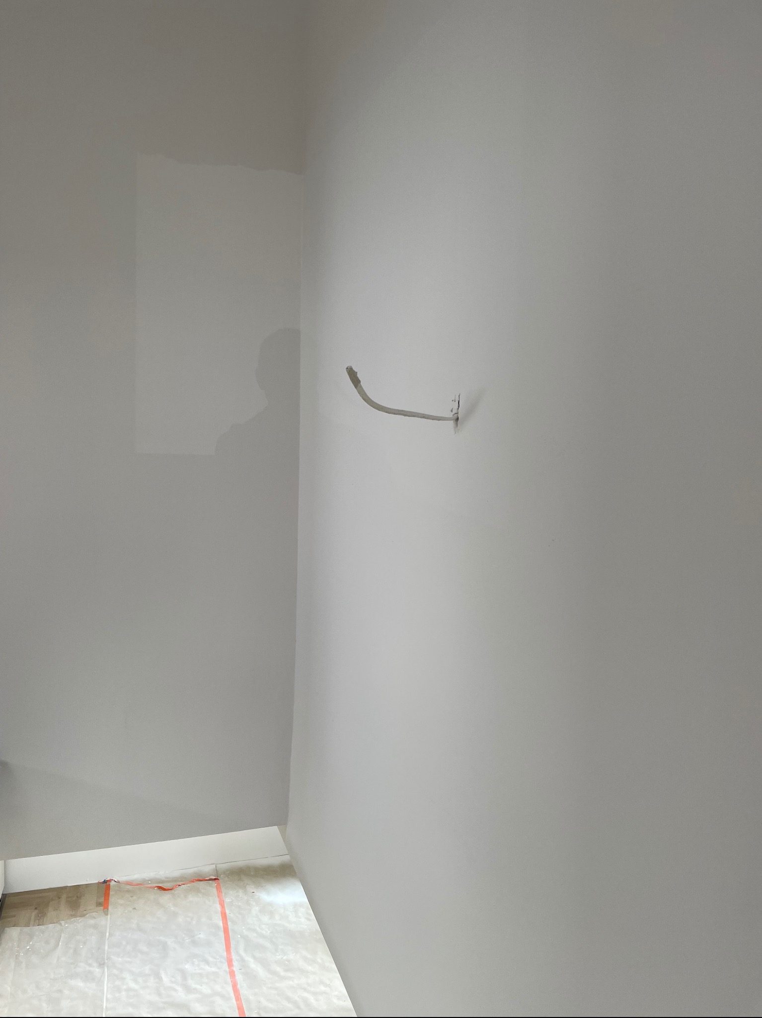 Gładka, jasnoszara ściana po tynkowaniu maszynowym z wystającym kablem elektrycznym. Podłoga zabezpieczona folią malarską. Widoczny cień osoby.