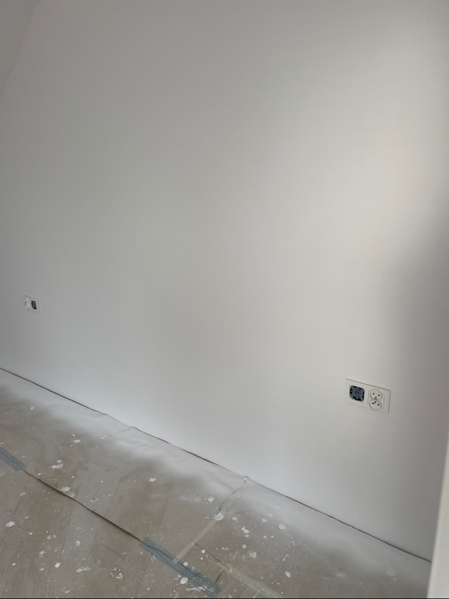 Gładka, biała ściana po tynkowaniu maszynowym z widocznymi gniazdkami elektrycznymi i zabezpieczoną podłogą. Minimalistyczne ujęcie wnętrza po remoncie.