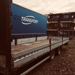 Ciężar&oacute;wka z otwartą plandeką i logo GTC Transport, zaparkowana na tle budynku mieszkalnego. Widok z boku, jesienna sceneria.