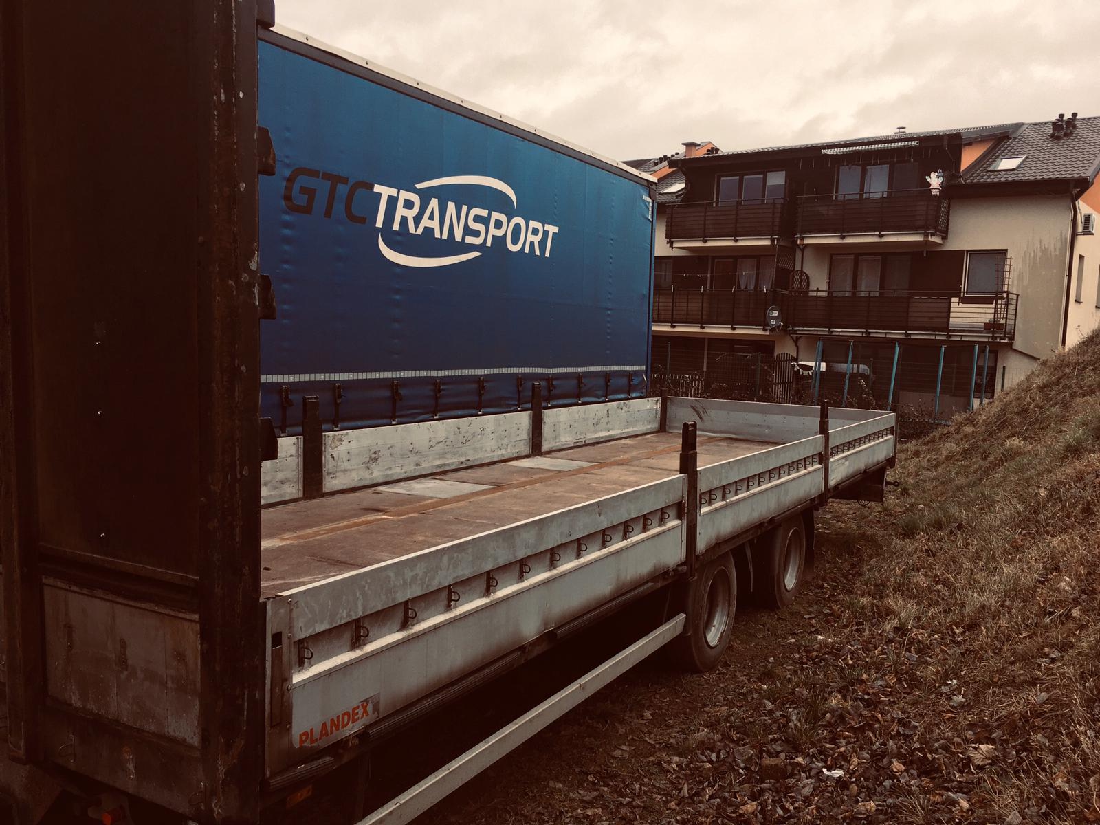 Ciężarówka z otwartą plandeką i logo GTC Transport, zaparkowana na tle budynku mieszkalnego. Widok z boku, jesienna sceneria.