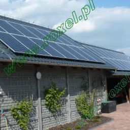 Panele słoneczne na dachu budynku z szarej dachówki, widoczne pnącza na siatce przy ścianie. Dach z zamontowaną instalacją fotowoltaiczną w słoneczny dzień.