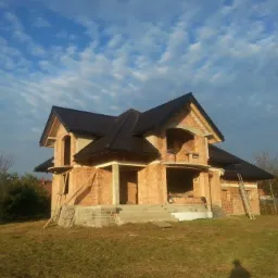 Niedokończony dwukondygnacyjny dom z czerwonej cegły z ciemnobrązowym dachem na tle błękitnego nieba z chmurami, widoczne drewniane drabiny i częściowe wykończenie elewacji.