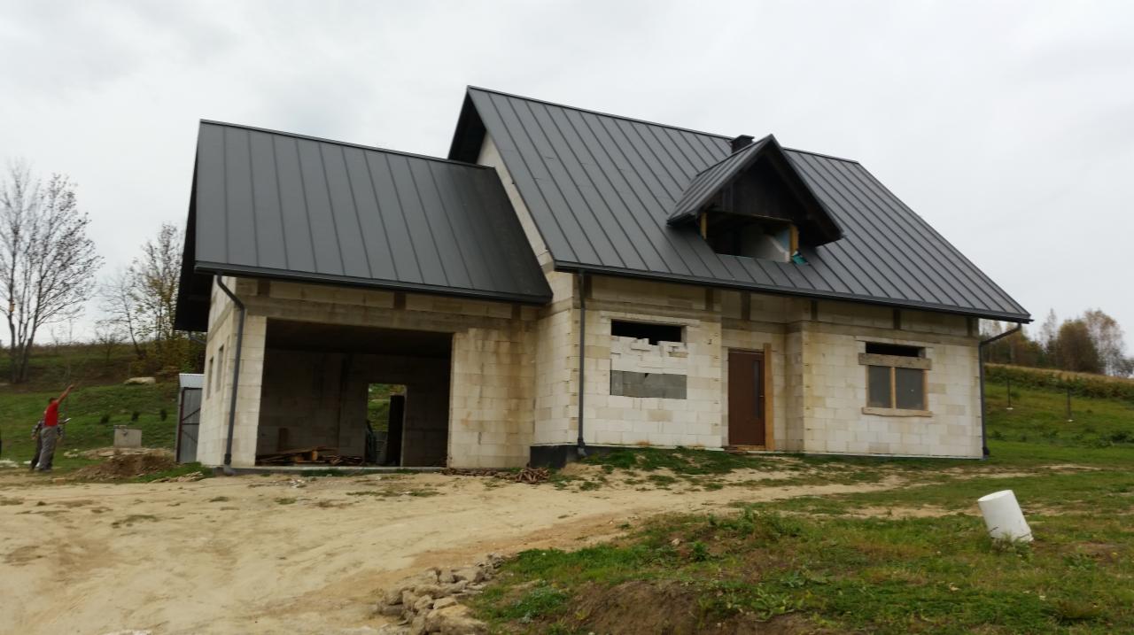 Surowy stan domu murowanego z ciemnym dachem z blachy, widoczny garaż, częściowo wykończona elewacja i pracownik na zewnątrz.