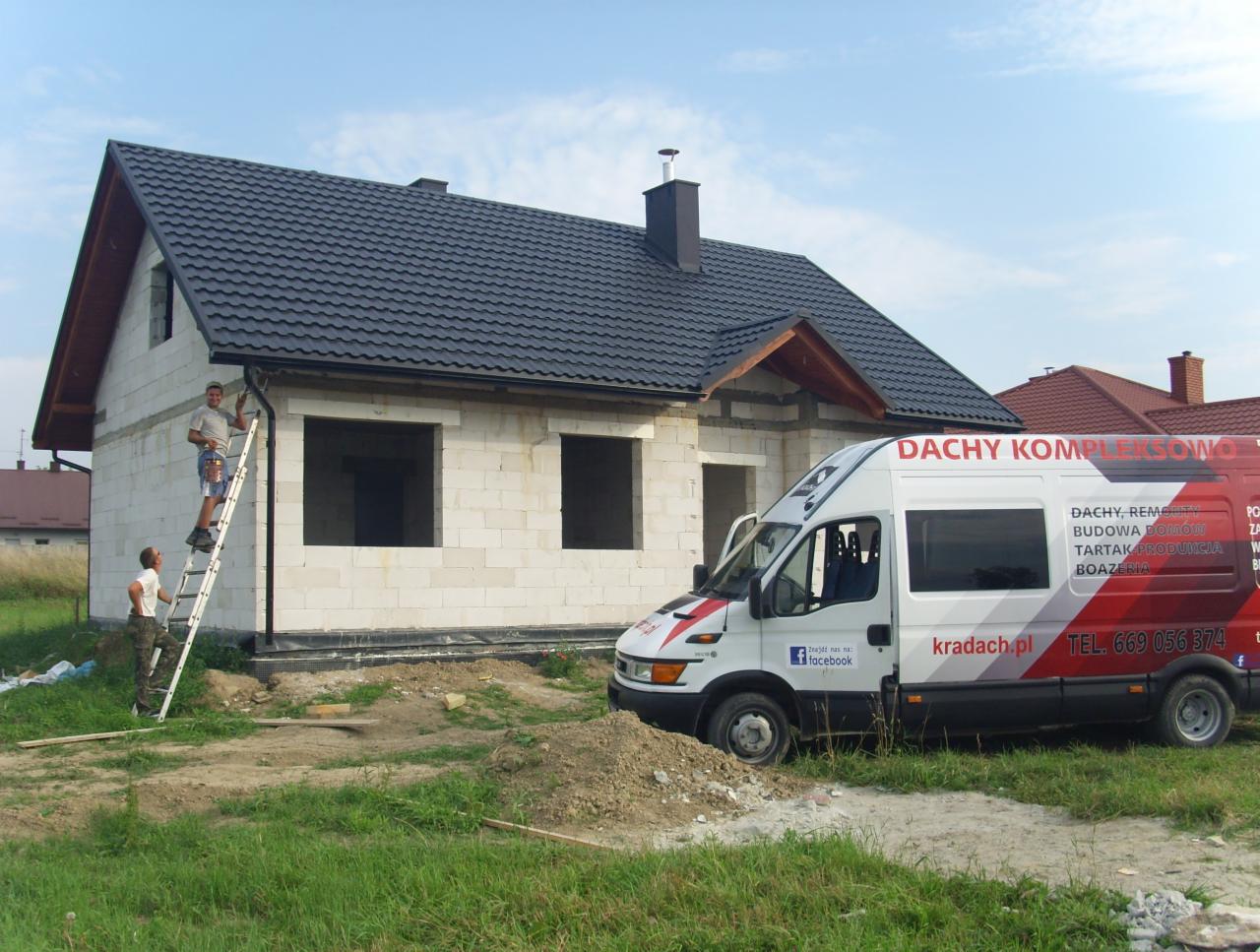 Budowa domu z białego bloczka w stanie surowym, z zamontowanym czarnym dachem z blachodachówki. Dwóch mężczyzn montuje rynny na drabinie. Na pierwszym planie samochód dostawczy z reklamą firmy...