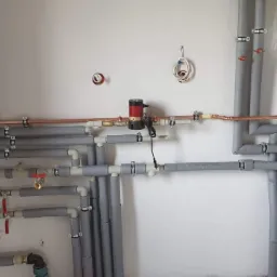 Nowa instalacja centralnego ogrzewania z rur miedzianych i szarych otulin, widoczny piec gazowy zabezpieczony folią, manometr i liczne zawory na białej ścianie.