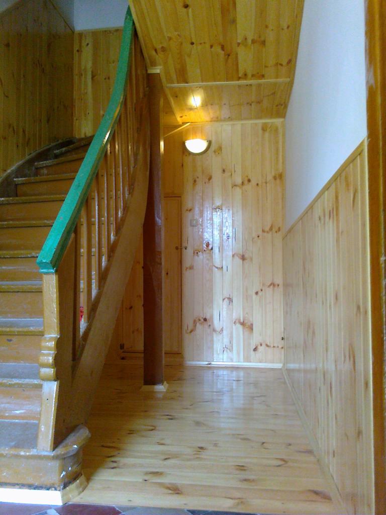 Korytarz wykończony boazerią drewnianą, z widocznymi schodami z zieloną barierką, oświetleniem sufitowym i błyszczącą podłogą.