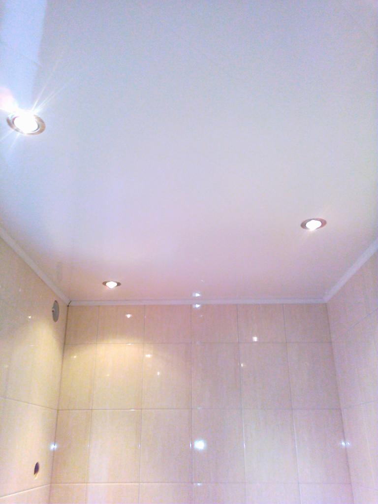 Wykończona łazienka z beżowymi płytkami, podwieszanym sufitem i trzema punktowymi lampami halogenowymi. Widoczne otwory na instalacje elektryczne w ścianie.
