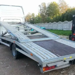 Pusta laweta z rozłożonymi najazdami, obok wózek widłowy Linde, w tle betonowy płot i drzewa.