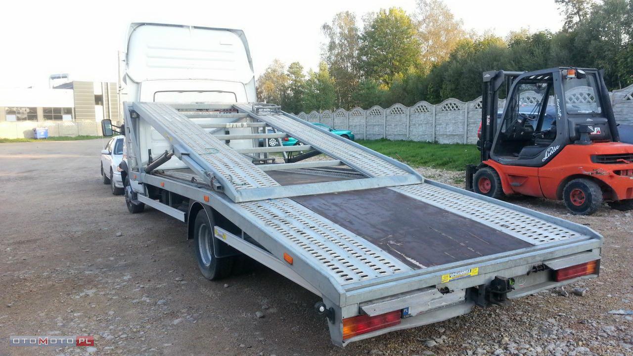 Pusta laweta z rozłożonymi najazdami, obok wózek widłowy Linde, w tle betonowy płot i drzewa.