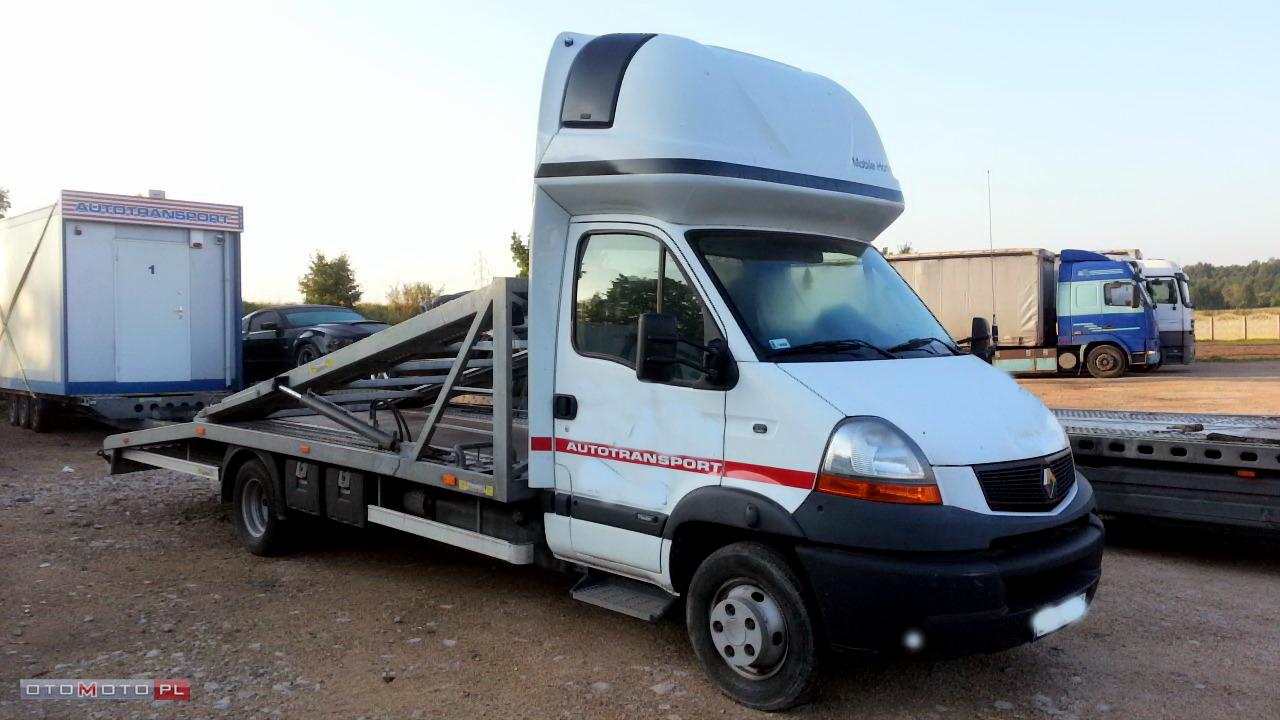 Biała laweta z napisem AUTOTRANSPORT i czerwoną linią, stojąca na żwirowym placu, ciągnąca białą przyczepę-kontener. W tle zaparkowane inne ciężarówki i drzewa.