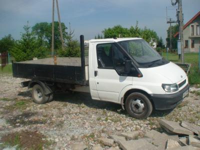 Biały samochód ciężarowy Ford Transit wywrotka do 3.5t z czarną skrzynią ładunkową, zaparkowany na żwirowym podłożu, w tle dom jednorodzinny i słup energetyczny.