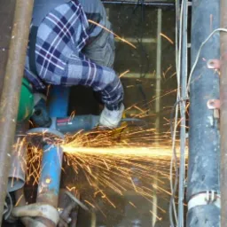 Pracownik w kasku i okularach ochronnych przecina metalową rurę szlifierką kątową, iskry rozbryzgują się dookoła.