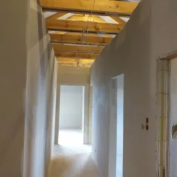 Wnętrze domu w trakcie wykańczania: świeżo otynkowane ściany w odcieniach szarości, surowa drewniana konstrukcja dachu z widocznymi kablami, perspektywa korytarza z otworami drzwiowymi.