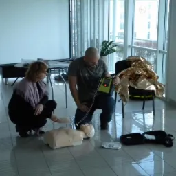 Instruktor demonstruje użycie defibrylatora AED na fantomie, podczas gdy kursantka ćwiczy resuscytację na manekinie na podłodze w przestronnym pomieszczeniu z dużymi oknami.