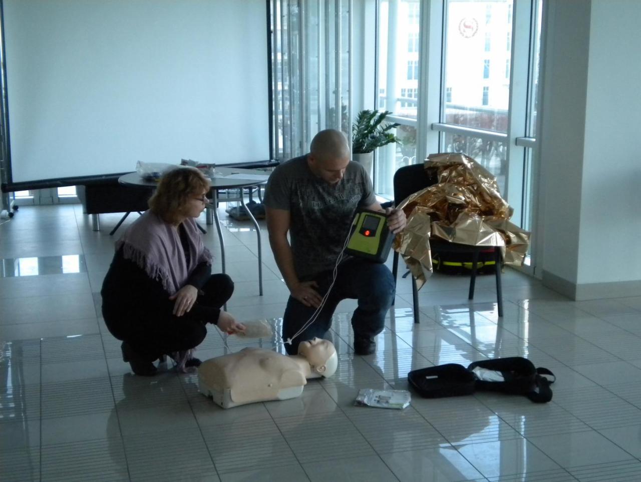 Instruktor demonstruje użycie defibrylatora AED na fantomie, podczas gdy kursantka ćwiczy resuscytację na manekinie na podłodze w przestronnym pomieszczeniu z dużymi oknami.