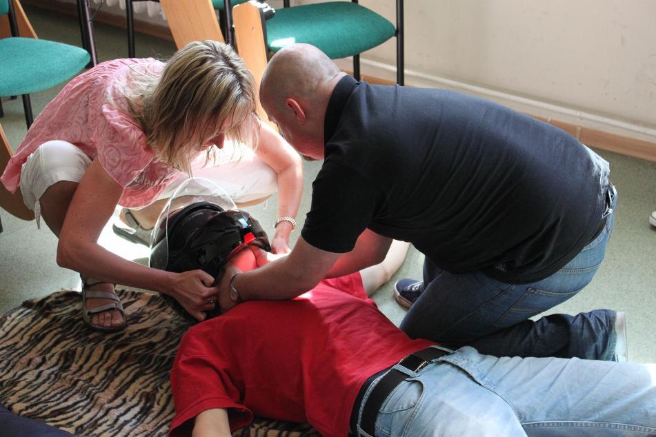 Osoby ćwiczą udzielanie pomocy poszkodowanemu w kasku na tle zielonych krzeseł i dywanu w zebrę.