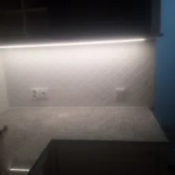 Białe płytki o marokańskim wzorze na ścianie nad szarym blatem kuchennym, oświetlone listwą LED, widoczne gniazdka elektryczne i włącznik.