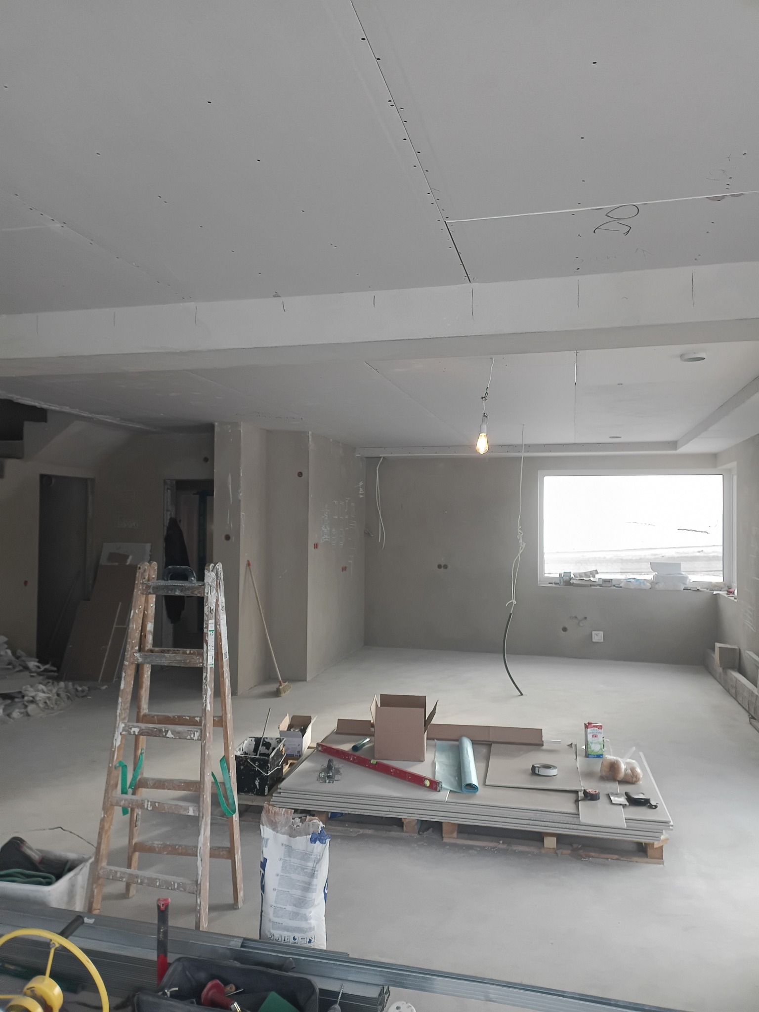 Wnętrze w trakcie montażu płyt karton-gips na suficie, widoczne narzędzia, drabina i materiały budowlane. Surowy stan wykończenia, przygotowanie do dalszych prac.