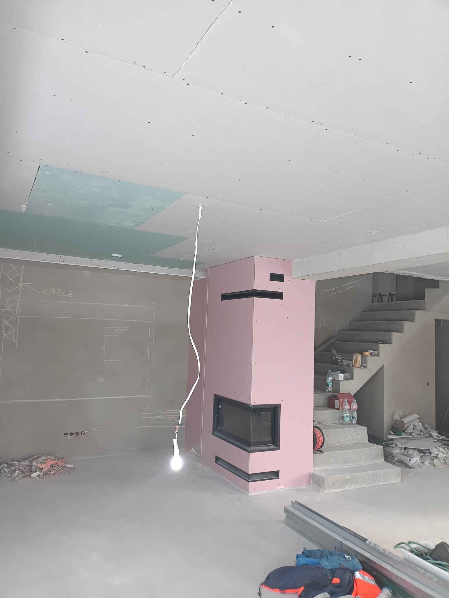 Wnętrze w trakcie wykończenia: różowy kominek obudowany płytami GK, sufit z częściowo zamontowanymi płytami, widoczne schody i prowizoryczne oświetlenie.