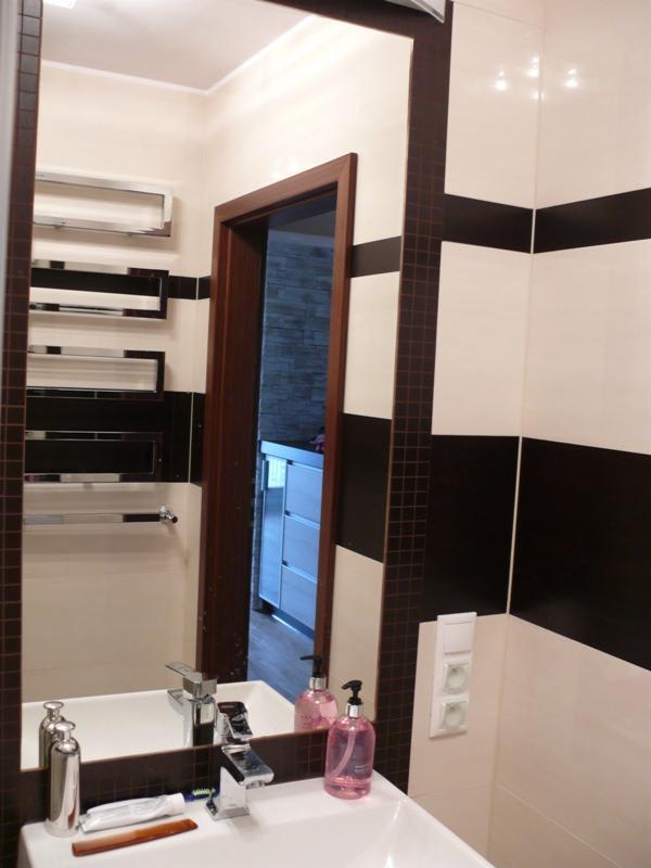 Wykończona łazienka z poziomymi pasami czarno-białych płytek, widoczna w lustrze odbijającym umywalkę z dozownikami mydła i fragmentem otwartych drzwi do innego pomieszczenia.