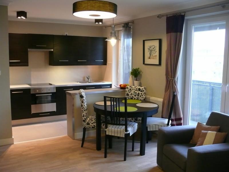 Nowoczesne wnętrze mieszkania z aneksem kuchennym, czarne szafki kuchenne z metalowymi uchwytami, okrągły stół z krzesłami o różnym wzornictwie, szary fotel z poduszkami w paski, jasna drewniana...