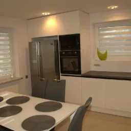 Nowoczesna kuchnia z białymi szafkami, srebrną lodówką i czarnym piekarnikiem. Na pierwszym planie biały stół z szarymi podkładkami i krzesłami.