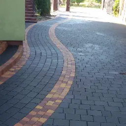 Zakrzywiony podjazd z ciemnoszarej kostki brukowej z dekoracyjnym pasem z kostki w odcieniach żółci i czerwieni, prowadzący od schodów wejściowych do bramy wjazdowej.