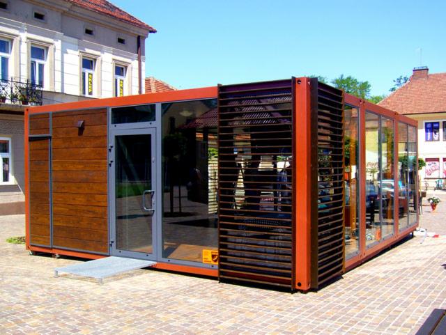Gotowa konstrukcja modułowa pawilonu handlowego z drewnianą fasadą i przeszklonymi ścianami, z rampą wejściową, na tle miejskiego placu.
