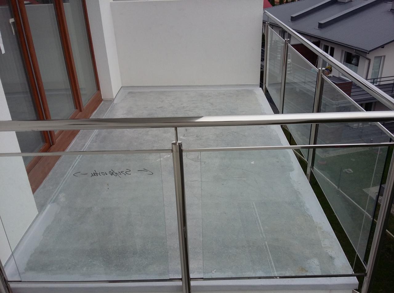 Balkon z betonową posadzką i szklanymi panelami w stalowej ramie, widoczne odręczne notatki na posadzce.