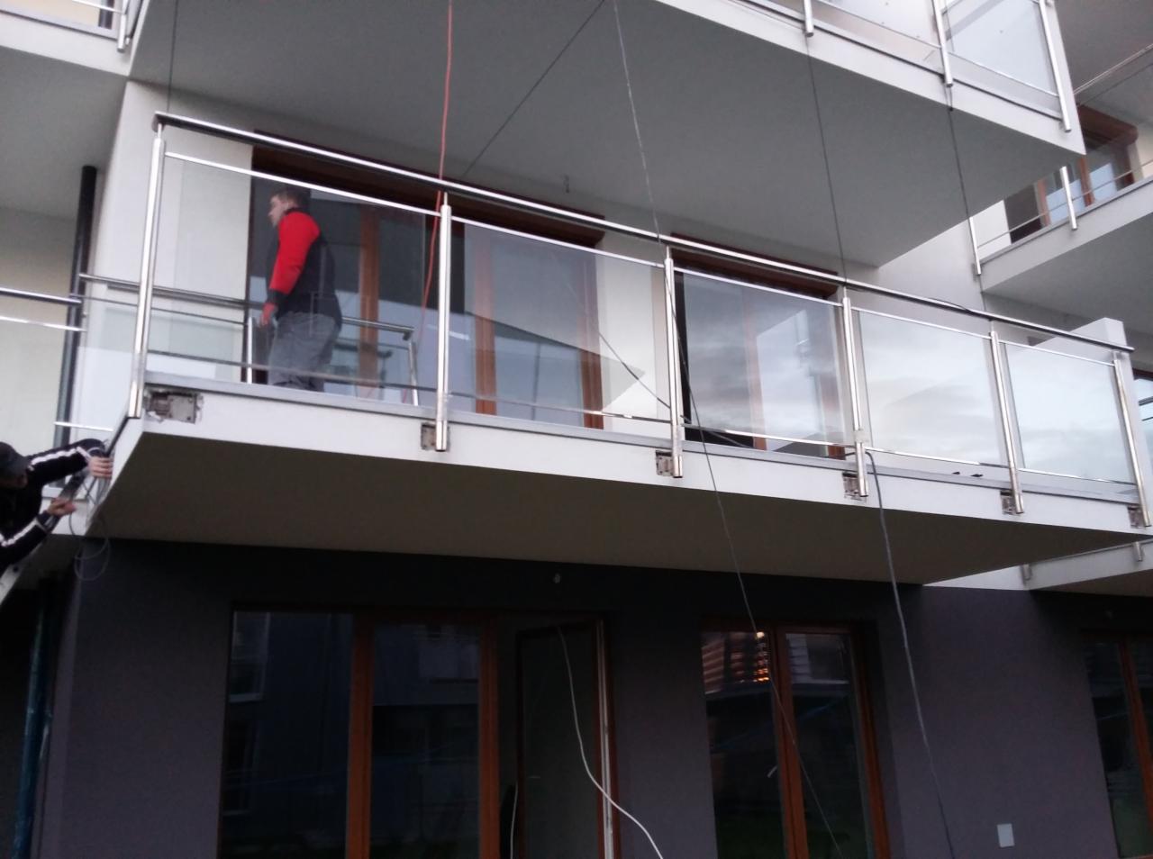 Montaż balustrady nierdzewnej ze szklanym wypełnieniem na balkonie budynku wielorodzinnego; widoczni dwaj monterzy podczas pracy.