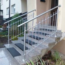 Zewnętrzne schody granitowe z metalową balustradą o prostym wzorze, zakończoną dekoracyjnymi elementami, prowadzące do wejścia budynku.