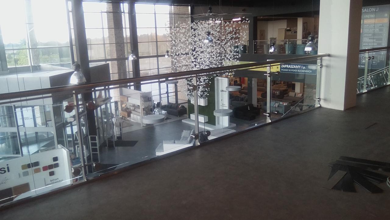 Wnętrze salonu meblowego z przeszkloną balustradą na antresoli, widok na ekspozycję mebli wypoczynkowych i dekoracyjną instalację z białych elementów.