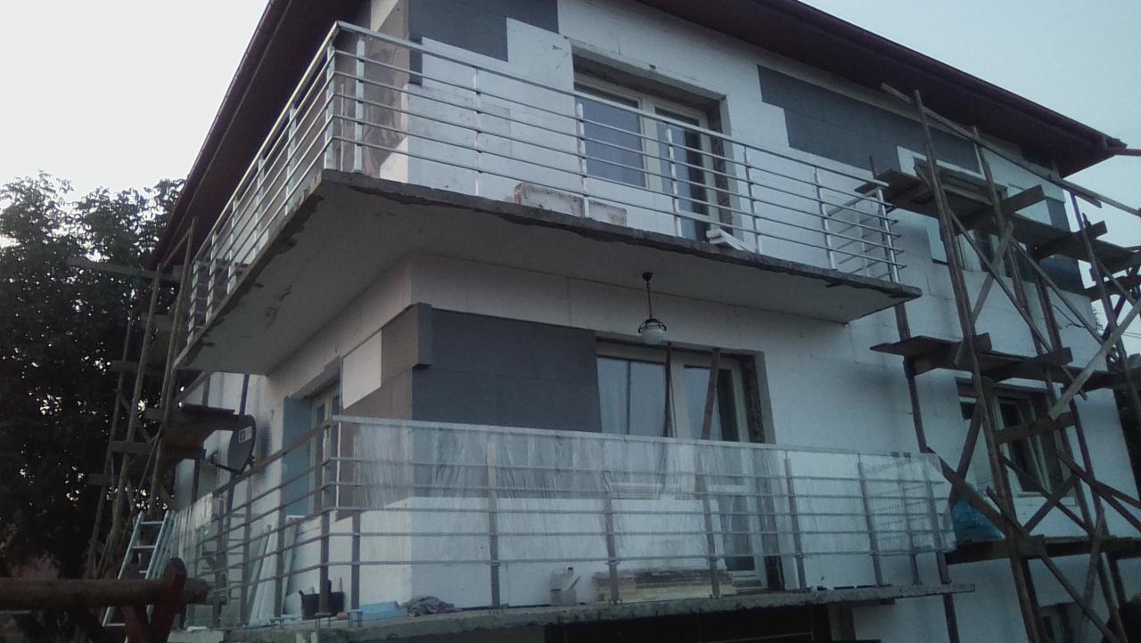 Budynek w trakcie wykończenia z zamontowanymi, poziomymi, metalowymi barierkami na balkonach, widoczne rusztowania i zabezpieczenia folią.