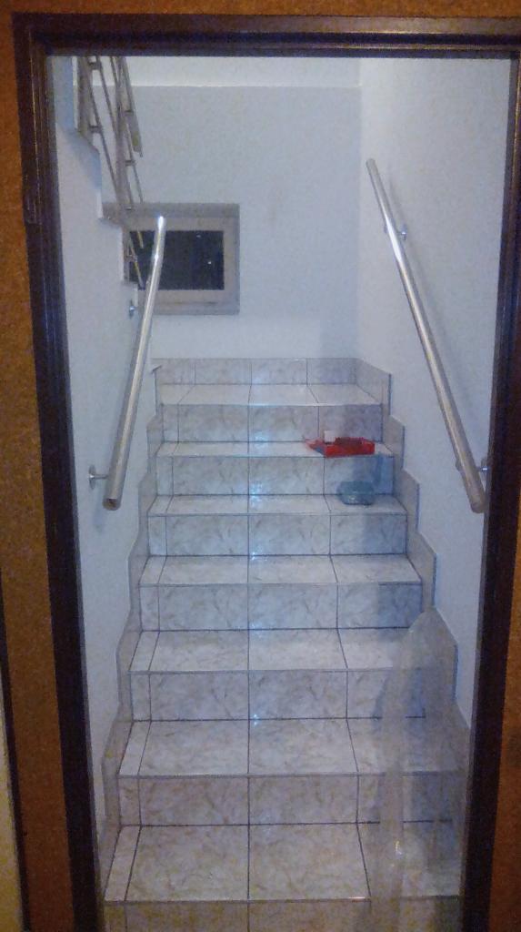 Wnętrze klatki schodowej z wykafelkowanymi schodami i metalowymi poręczami, widok z dołu przez ciemną framugę drzwi.