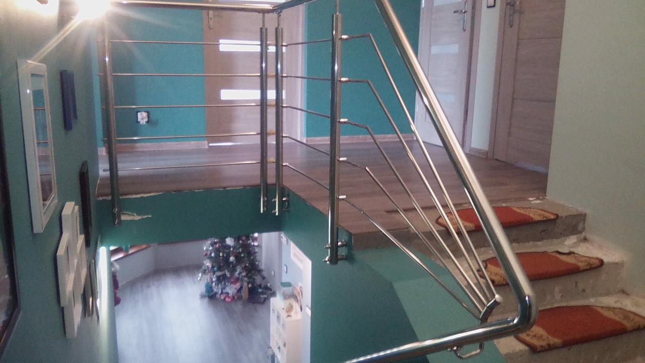 Wnętrze domu z widokiem na schody z metalową balustradą, na stopniach leżą małe dywaniki, w tle widoczna choinka.