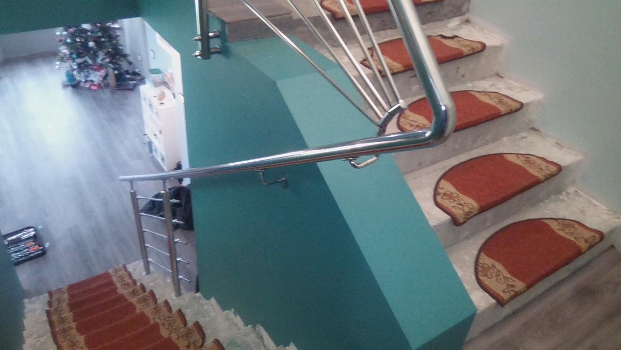 Schody z szarymi stopniami, czerwonymi nakładkami i metalową poręczą; widoczny fragment ściany w kolorze turkusowym i choinka w tle.