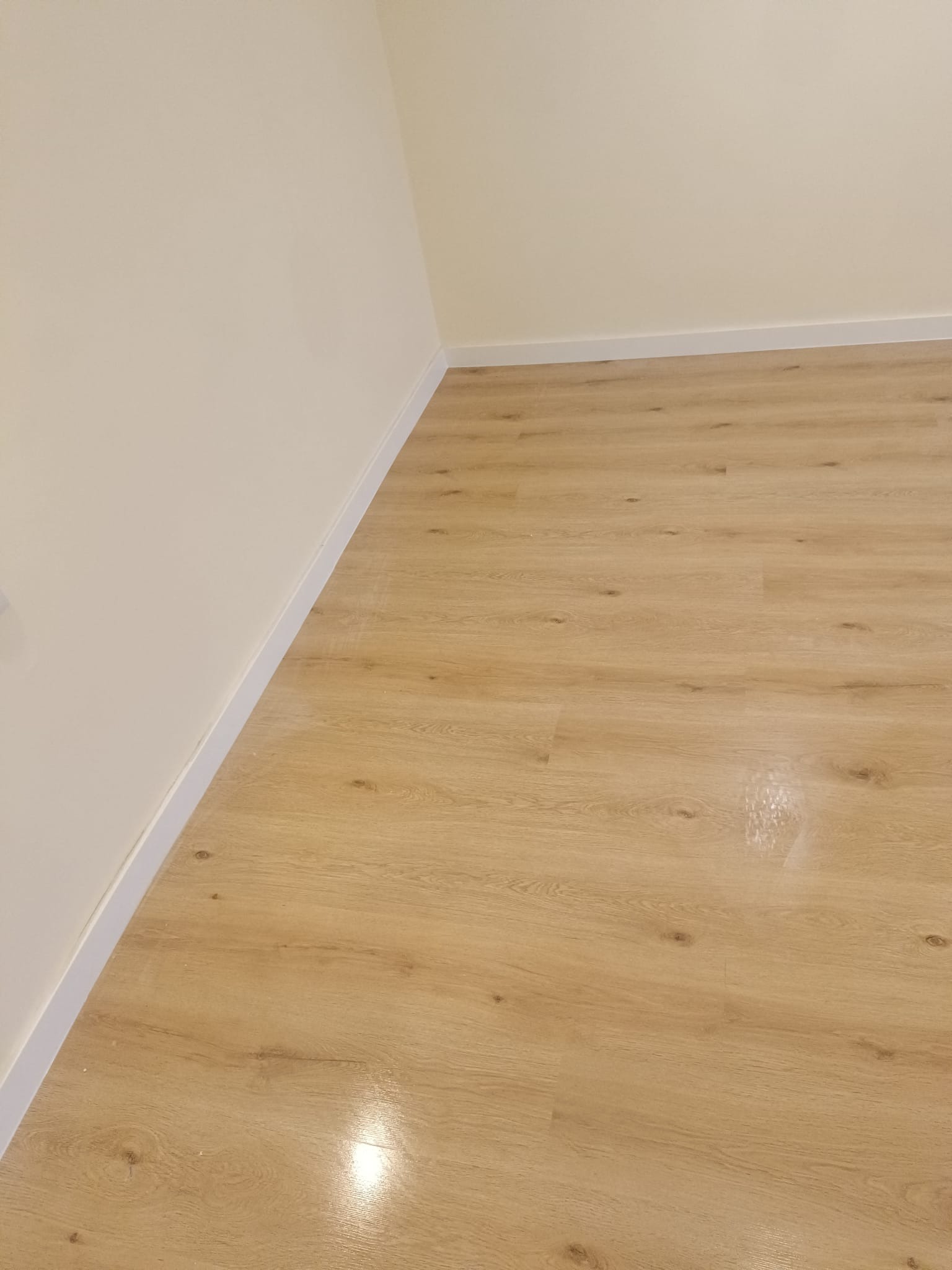 Jasne panele podłogowe z widoczną strukturą drewna, ułożone w rogu pomieszczenia z kremowymi ścianami i białymi listwami przypodłogowymi. Widok z góry.