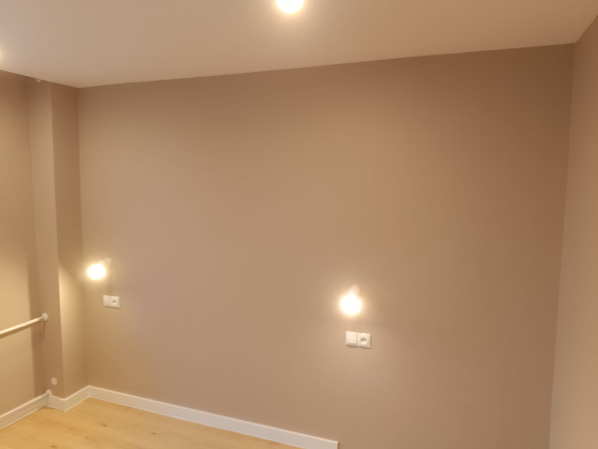 Beżowa ściana z dwoma kinkietami i gniazdkami, jasna podłoga z listwami przypodłogowymi. Minimalistyczne wnętrze po remoncie, oświetlenie punktowe.