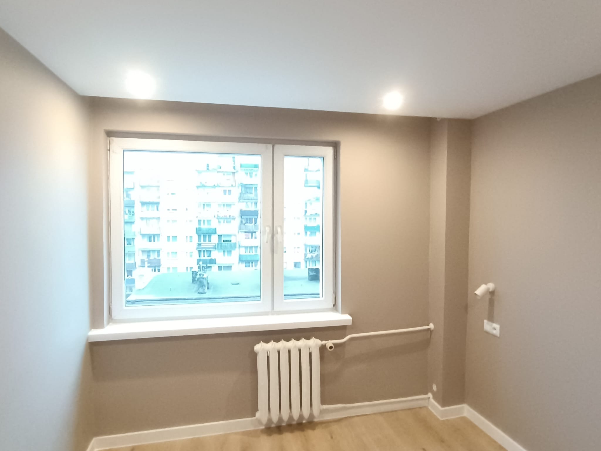 Jasne wnętrze po remoncie: beżowe ściany, białe okno z widokiem na blok, grzejnik i drewnopodobna podłoga. Minimalistyczny design z punktowym oświetleniem.