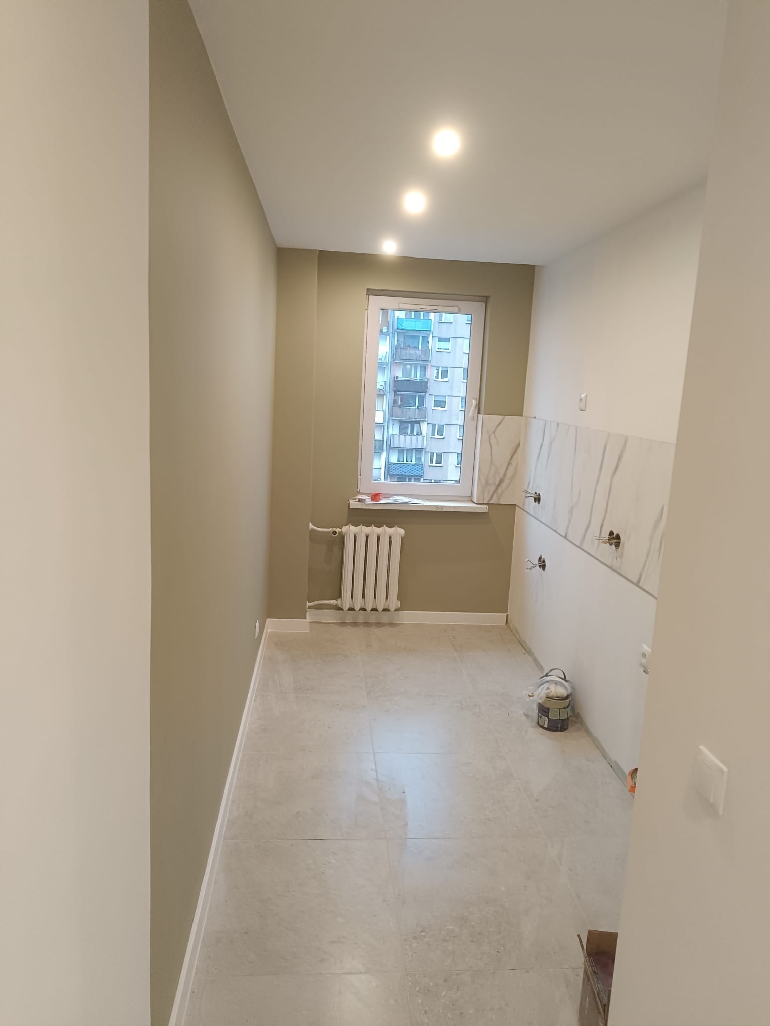 Wnętrze łazienki w trakcie prac wykończeniowych: pomalowane ściany w odcieniach beżu, częściowo ułożone płytki imitujące marmur, grzejnik pod oknem, puszka z farbą.