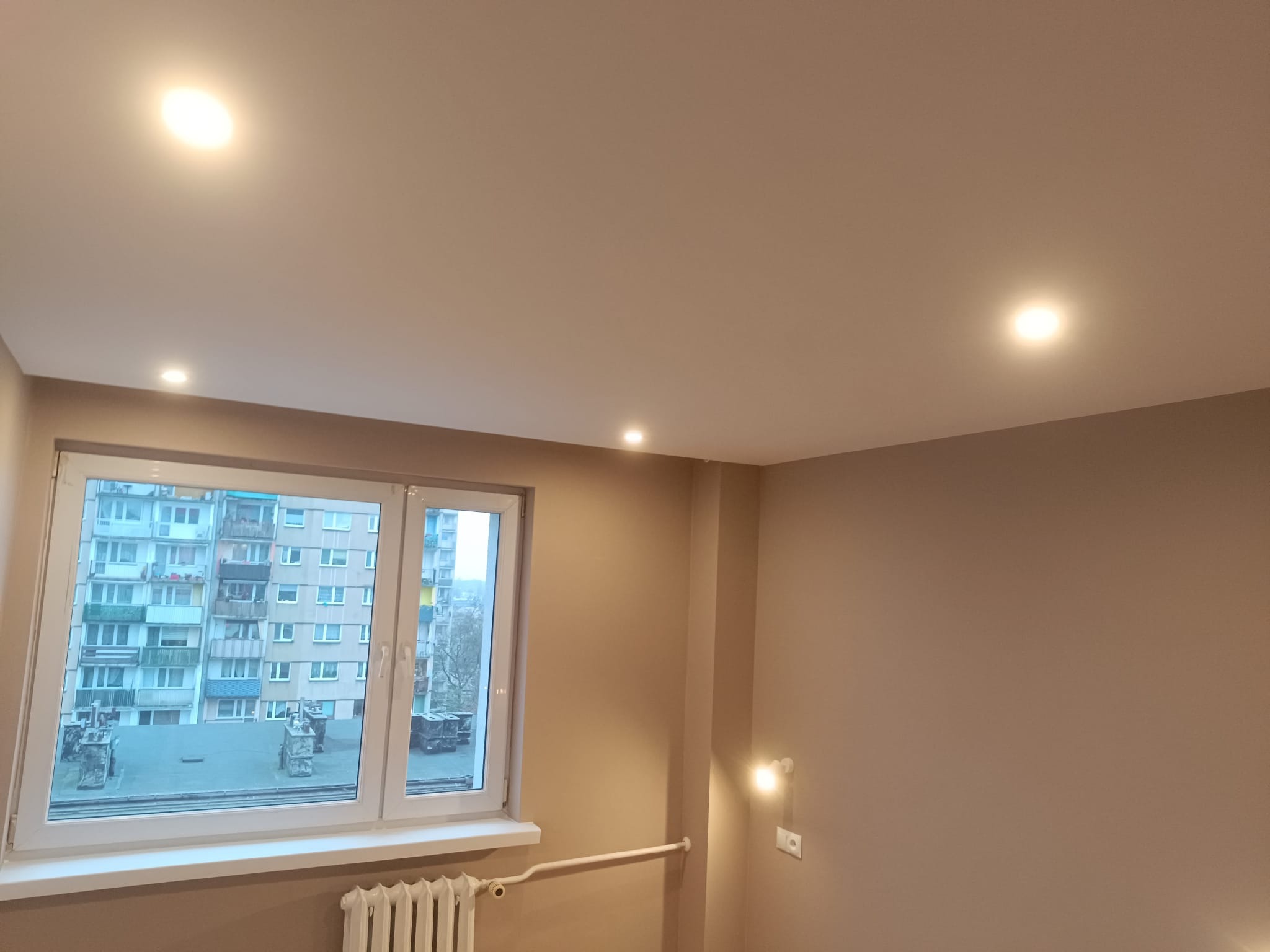 Jasne wnętrze po remoncie: gładki, beżowy sufit z trzema punktowymi lampami LED, ściany w odcieniach beżu, okno z widokiem na blok mieszkalny i biały grzejnik.