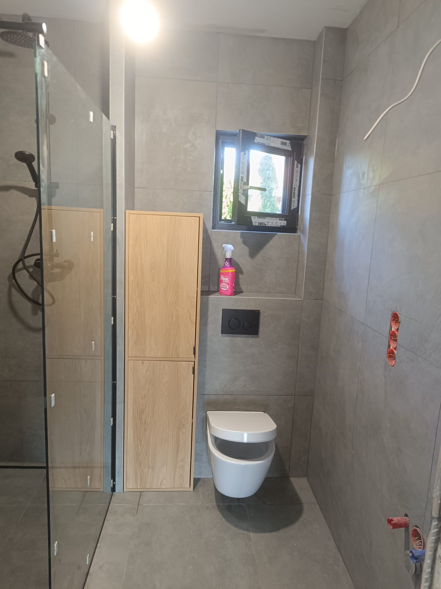 Nowoczesna łazienka w szarościach z kabiną prysznicową typu walk-in, wiszącą toaletą i szafką z drewnianym frontem. Widoczne detale wykończeniowe i okno.